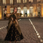 "Woman in a gold coat and black dress takes a selfie with her phone at night in a candlelit courtyard outside an elegant, well-lit building with large windows and cobblestone ground."